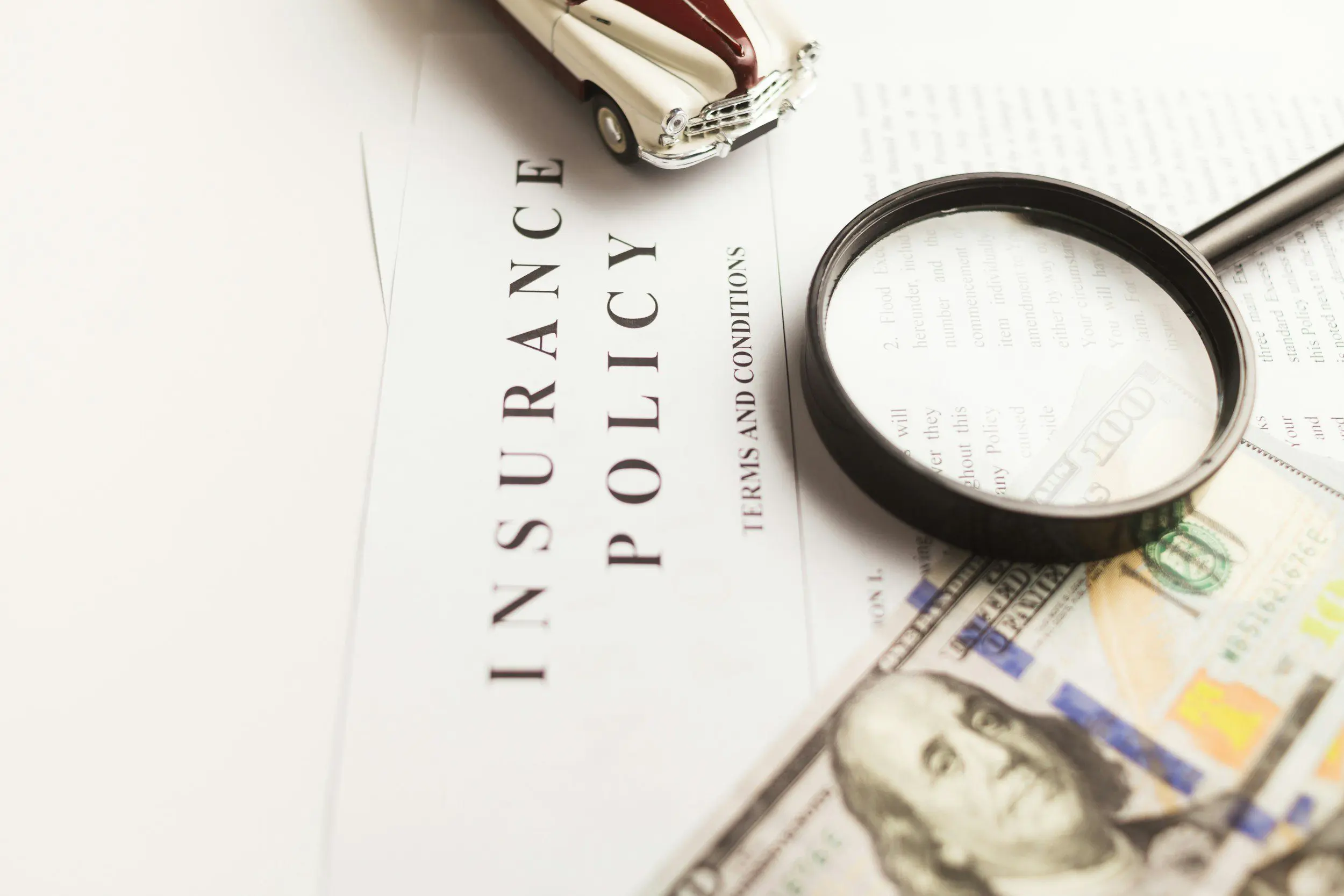 Magnifying glass and toy car on insurance policy document with US dollar bill.
