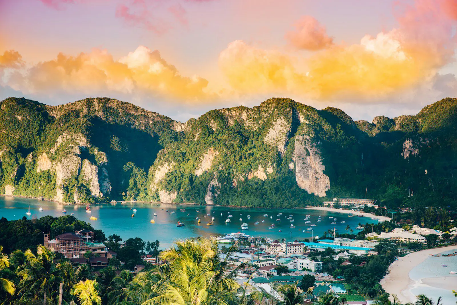 Aerial view of a tropical bay in Thailand surrounded by limestone cliffs, turquoise water, and colorful boats.