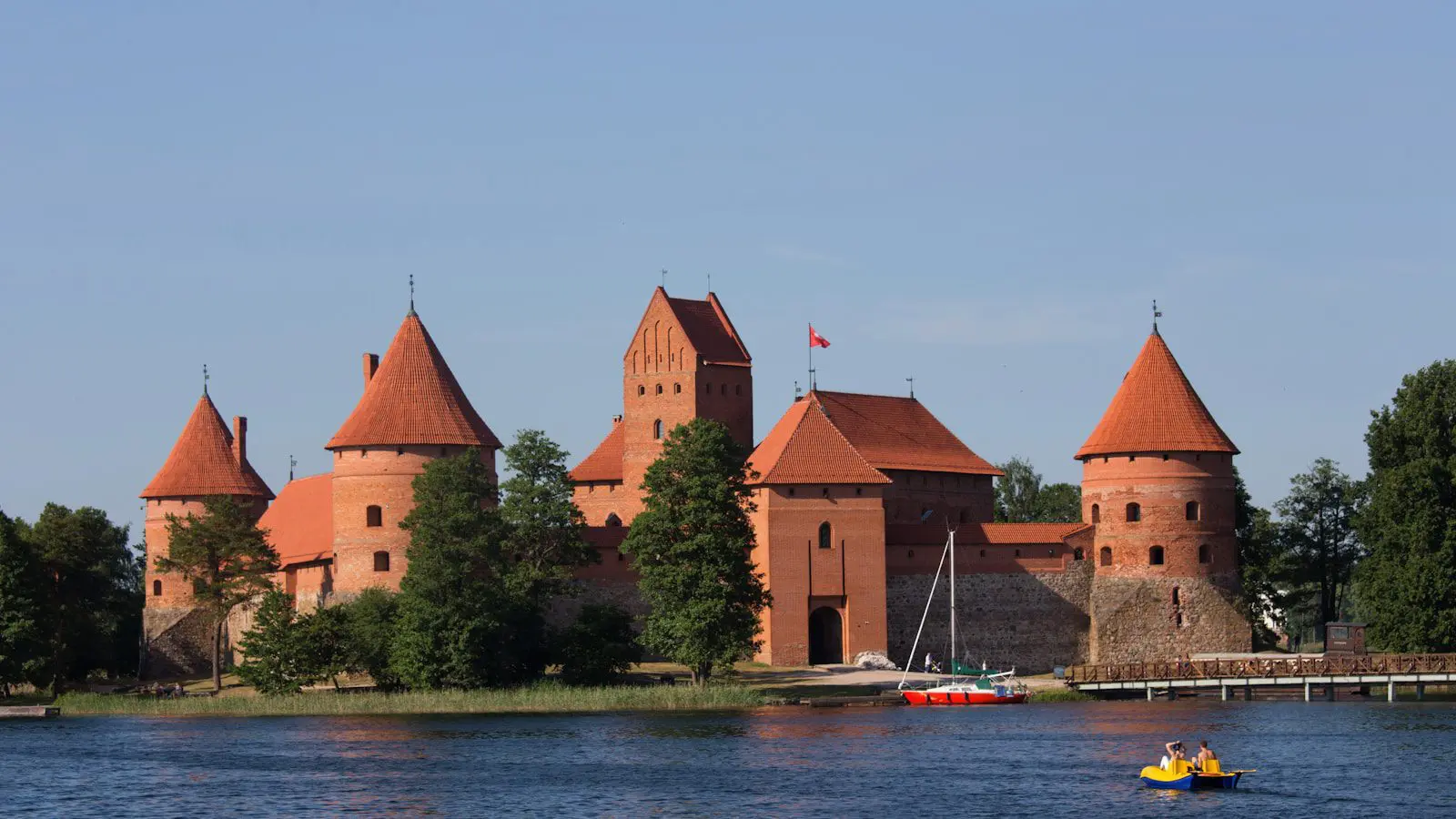 Photo by Gregory Atkats Historic red-brick Trakai Castle on Galvė Lake in Lithuania with boats on the water and blue summer sky.