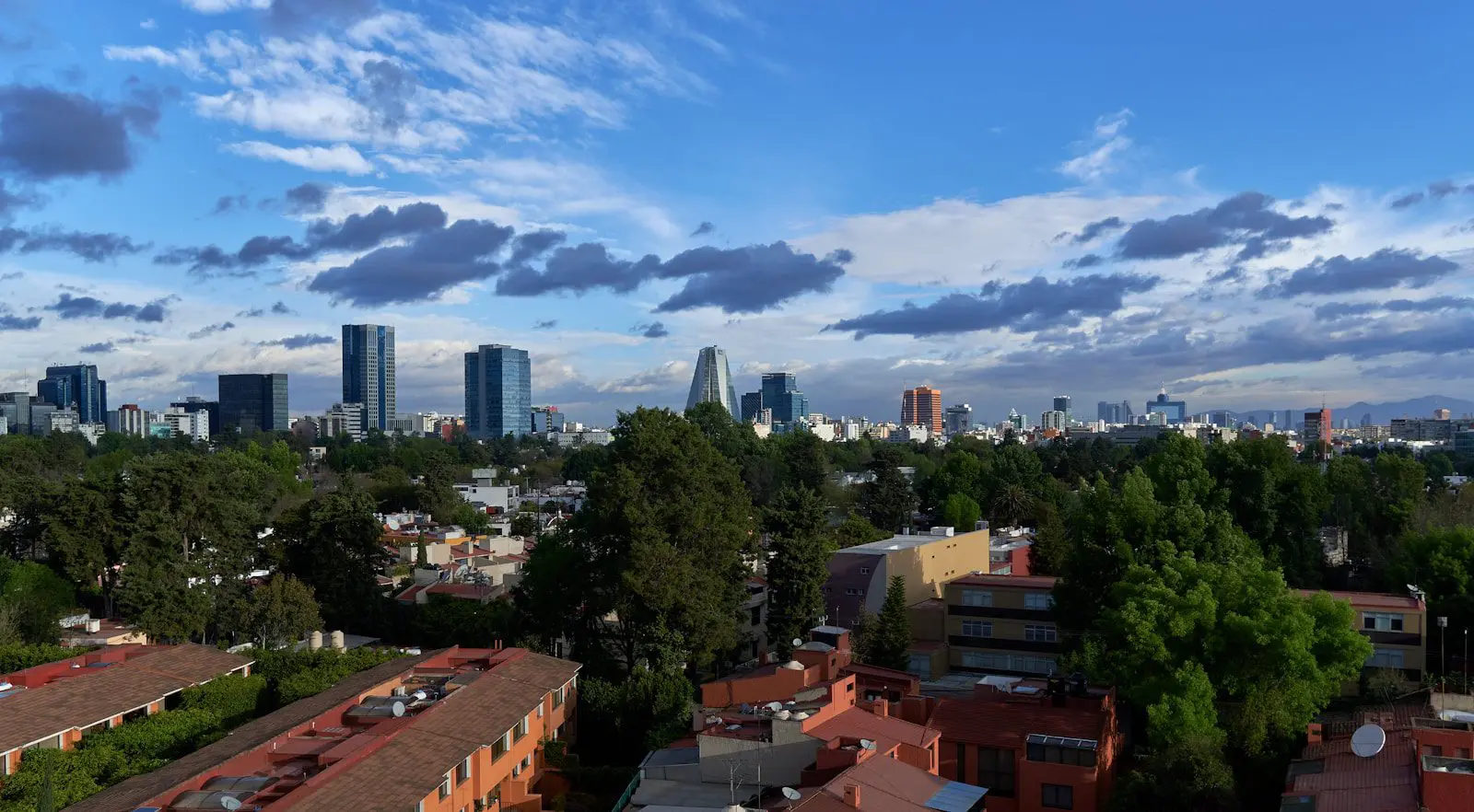 Panoramic view of Mexico City skyline with modern skyscrapers, trees, and blue sky with clouds.