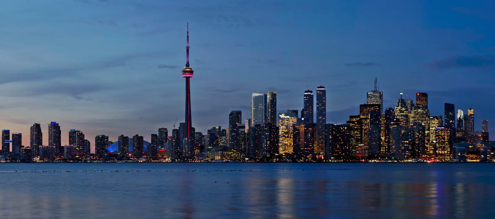 Canadian flag waving in the wind at dusk with city buildings and a soft pink sky in the background.