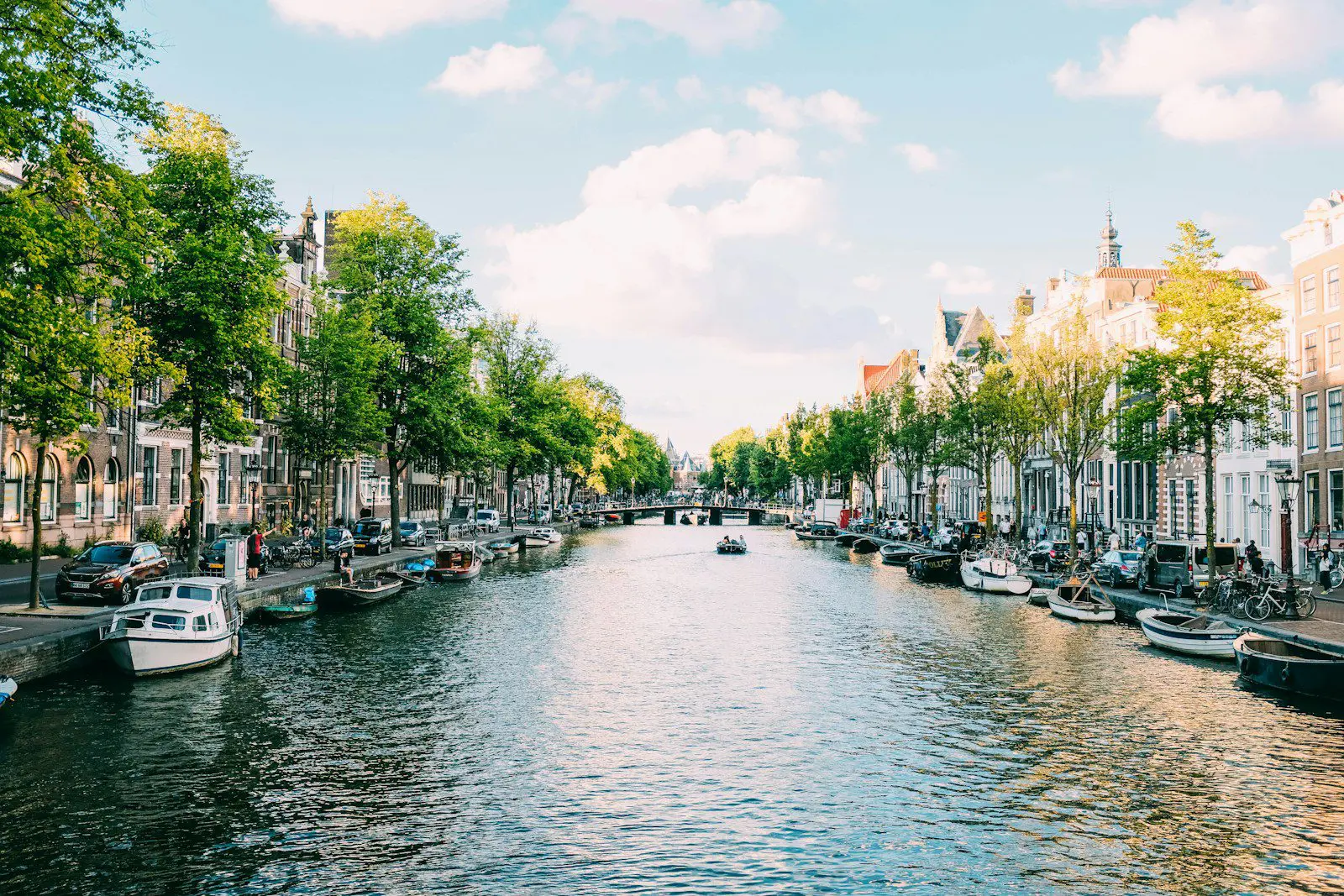 Scenic view of an Amsterdam canal lined with trees, boats, and traditional Dutch houses on a sunny day.