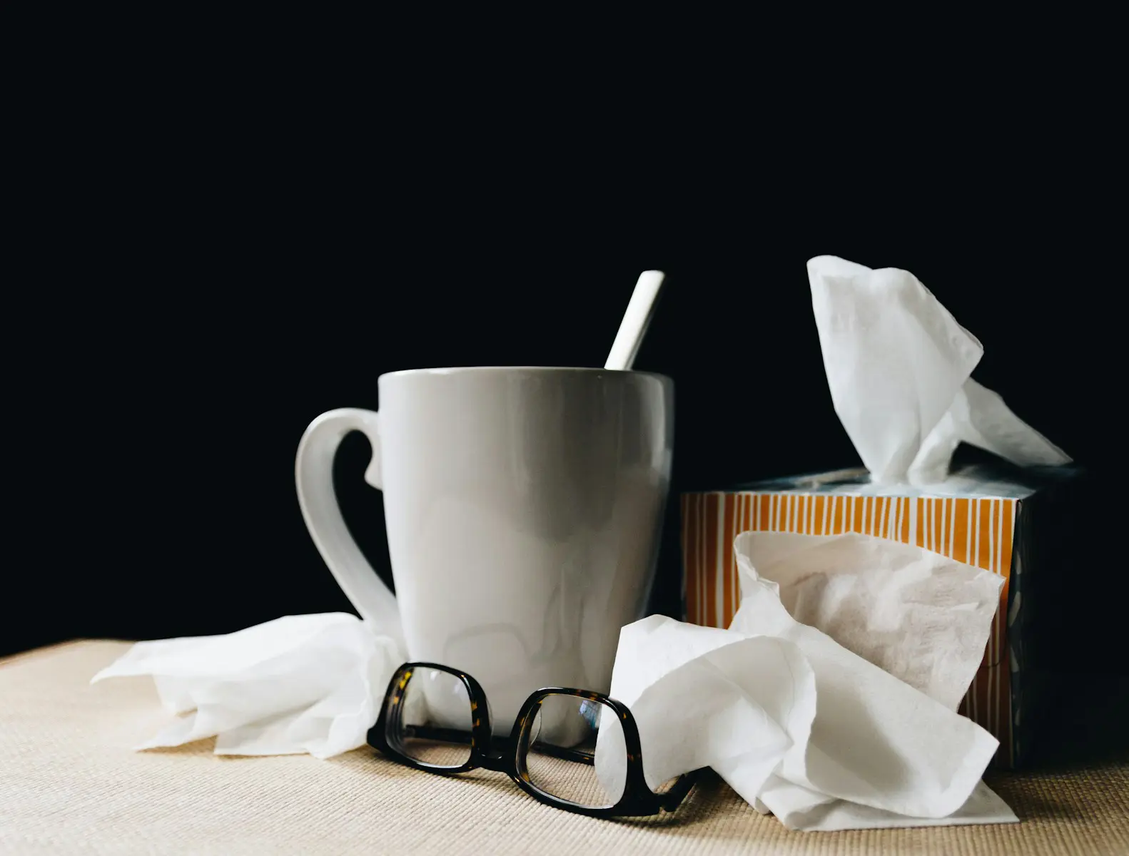 White mug, tissue box, and glasses on dark background, representing cold or flu season health care, home recovery, and self-care essentials.