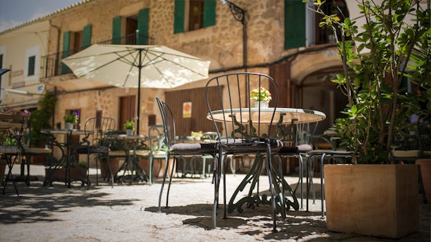 pexels-photo-1731826-1731826-1 A quaint outdoor café in Mallorca, Spain, with iron chairs and rustic stone buildings.